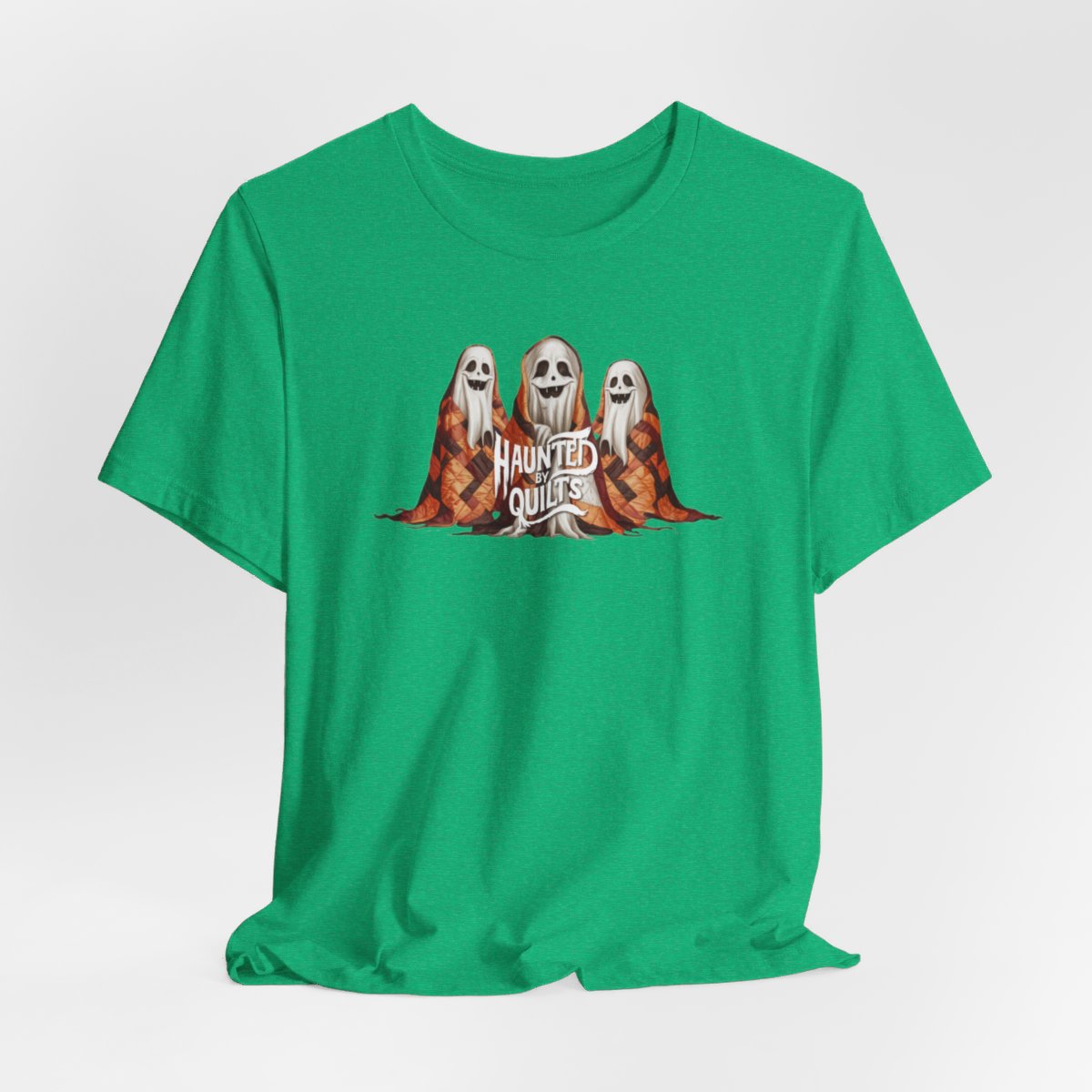 A Heather Kelly T-Shirt of three smiling cartoon ghosts covered in colorful patchwork quilts. The quilts feature orange, brown, and black patterns. Text below reads "HAUNTED BY QUILTS" in a spooky white font.