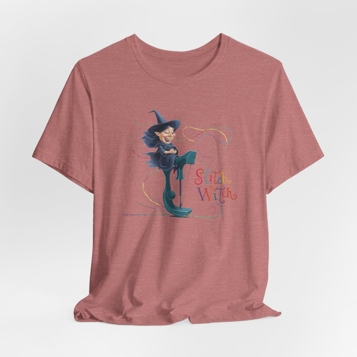 A Heather Mauve T-shirt with a cartoon illustration of a smiling witch with pointed hat riding a teal sewing machine. Colorful stitches swirl around her in the air. Text reads "Stitch Witch" in playful, multicolored letters.