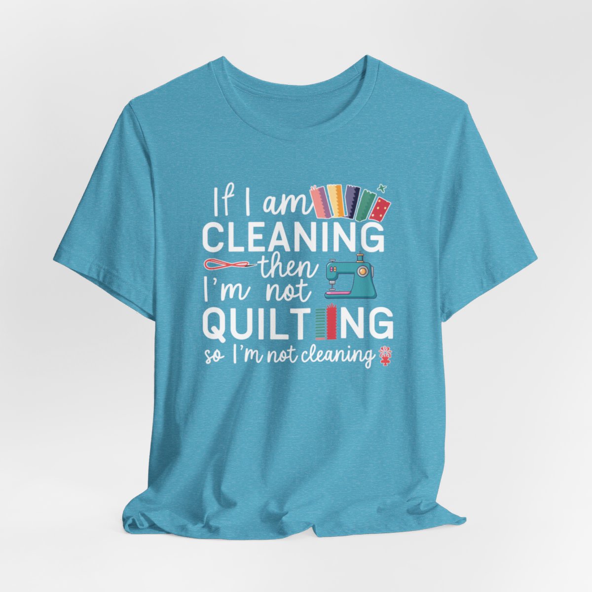 A Heather Aqua funny quilting T-Shirt with the phrase If I am Cleaning then I'm not Quilting in a bold and cheerful font with images of quilting icons and supplies