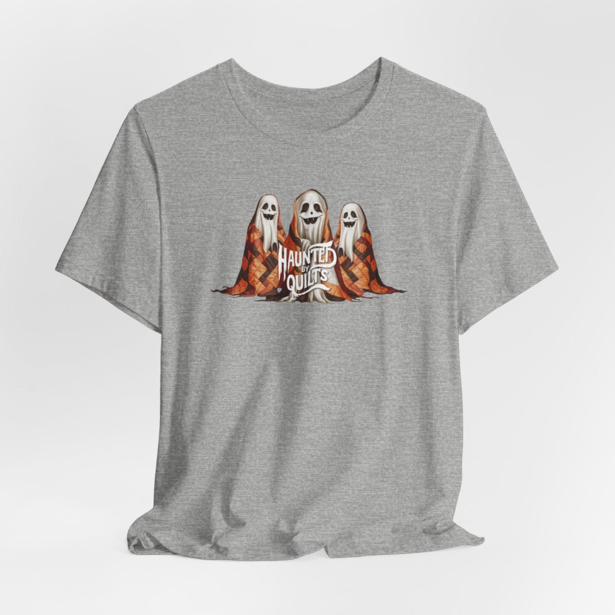 An Athletic Heather T-Shirt of three smiling cartoon ghosts covered in colorful patchwork quilts. The quilts feature orange, brown, and black patterns. Text below reads "HAUNTED BY QUILTS" in a spooky white font.