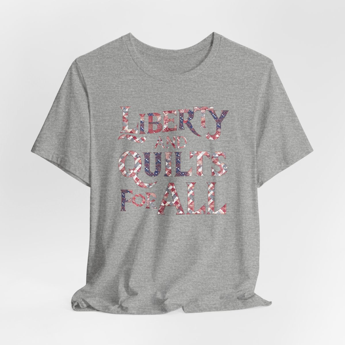 An Athletic Heather T-shirt featuring text "LIBERTY AND QUILTS FOR ALL" in large decorative letters filled with various quilt patterns in red, white, and blue.