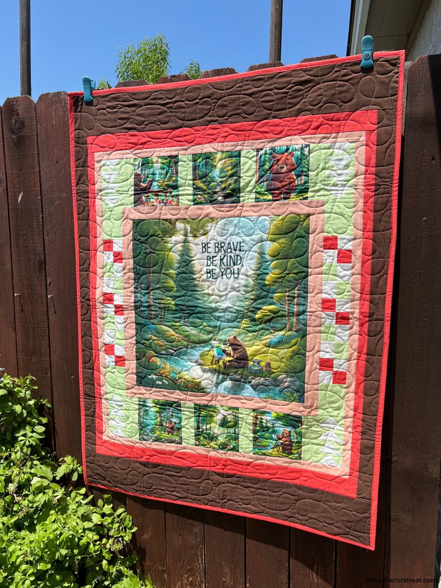 Be Kind Bear panel quilt kit finished in brown and coral, hung outdoors — bear-and-stream nursery quilt