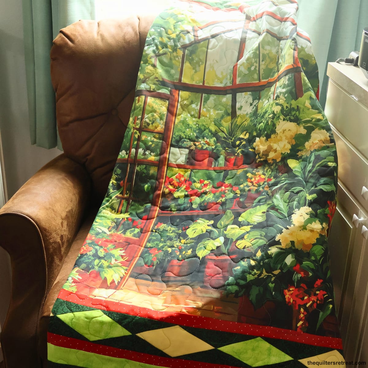 The Secret Greenhouse quilt draped on a chair — a detailed greenhouse panel quilt kit packed with potted plants