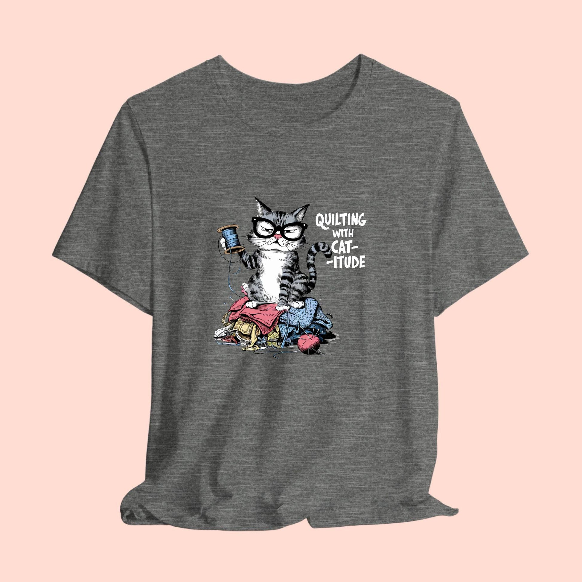 A t-shirt featuring a sassy cartoon cat sitting on a pile of colorful fabric, holding a spool of thread, with the phrase 'Quilting with Cat-itude,' perfect for quilters and cat lovers.