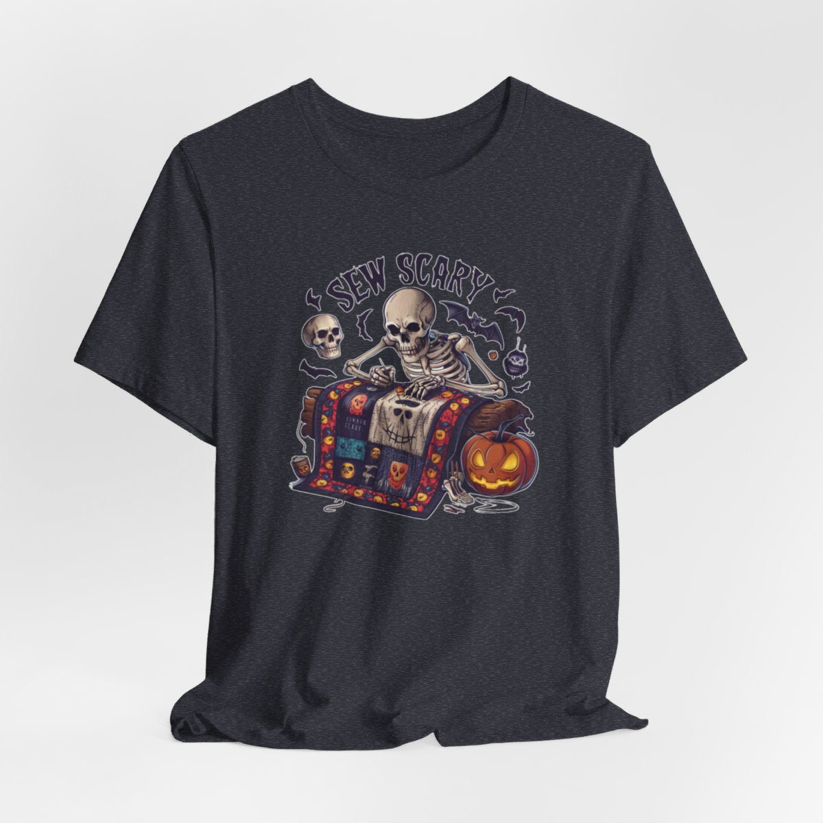 A Heather Navy Halloween Quilting T-Shirt featuring a skeleton sewing Halloween quilt. Colorful patchwork with spooky motifs. Bats and pumpkins surround skeleton. Text reads "Sew Scary". Ideal for quilters who love Halloween.