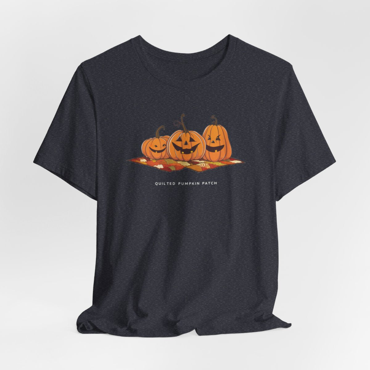 A Heather Navy T-shirt with three smiling jack-o'-lanterns sitting on a colorful autumn-themed quilt. The pumpkins have carved faces with various expressions, and the quilt beneath them features a patchwork of fall colors. Text below reads "QUILTED PUMPKIN PATCH".