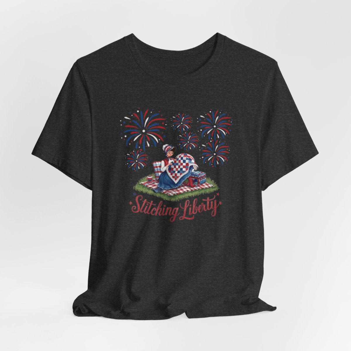 A Dark Grey Heather T-shirt with illustrated scene of a colonial woman quilting a patriotic blanket, surrounded by fireworks. Text reads "Stitching Liberty" in a decorative font.