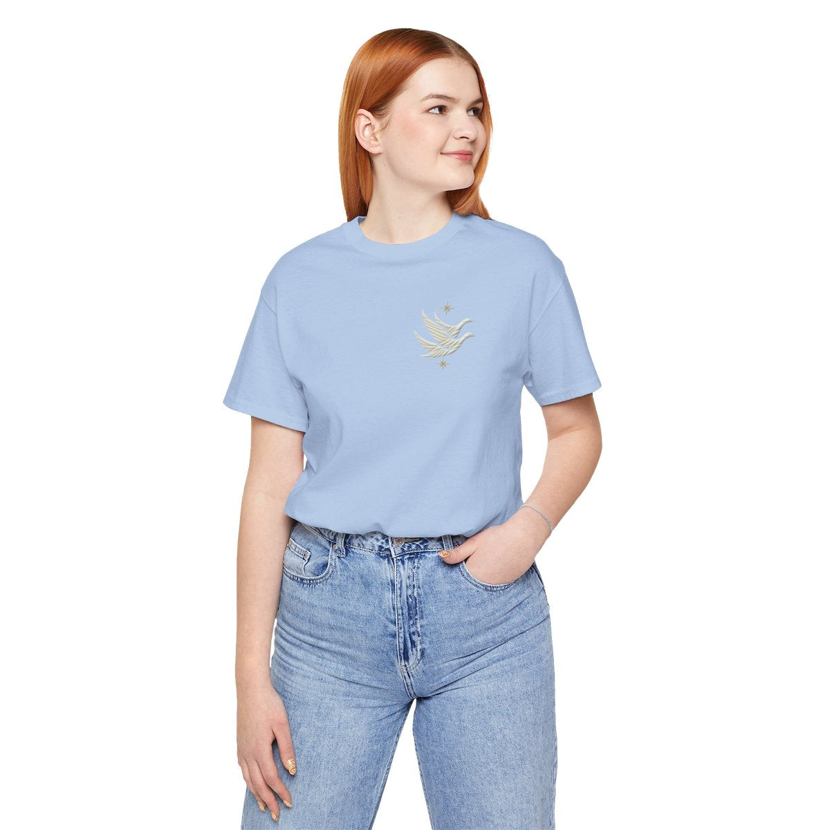 A Baby Blue T-shirt featuring two stylized white geese in flight, with outstretched wings. The geese are composed of intricate geometric patterns resembling quilt stitching. Two eight-pointed stars frame the scene, one above and one below the geese, adding a celestial element to the peaceful composition.
