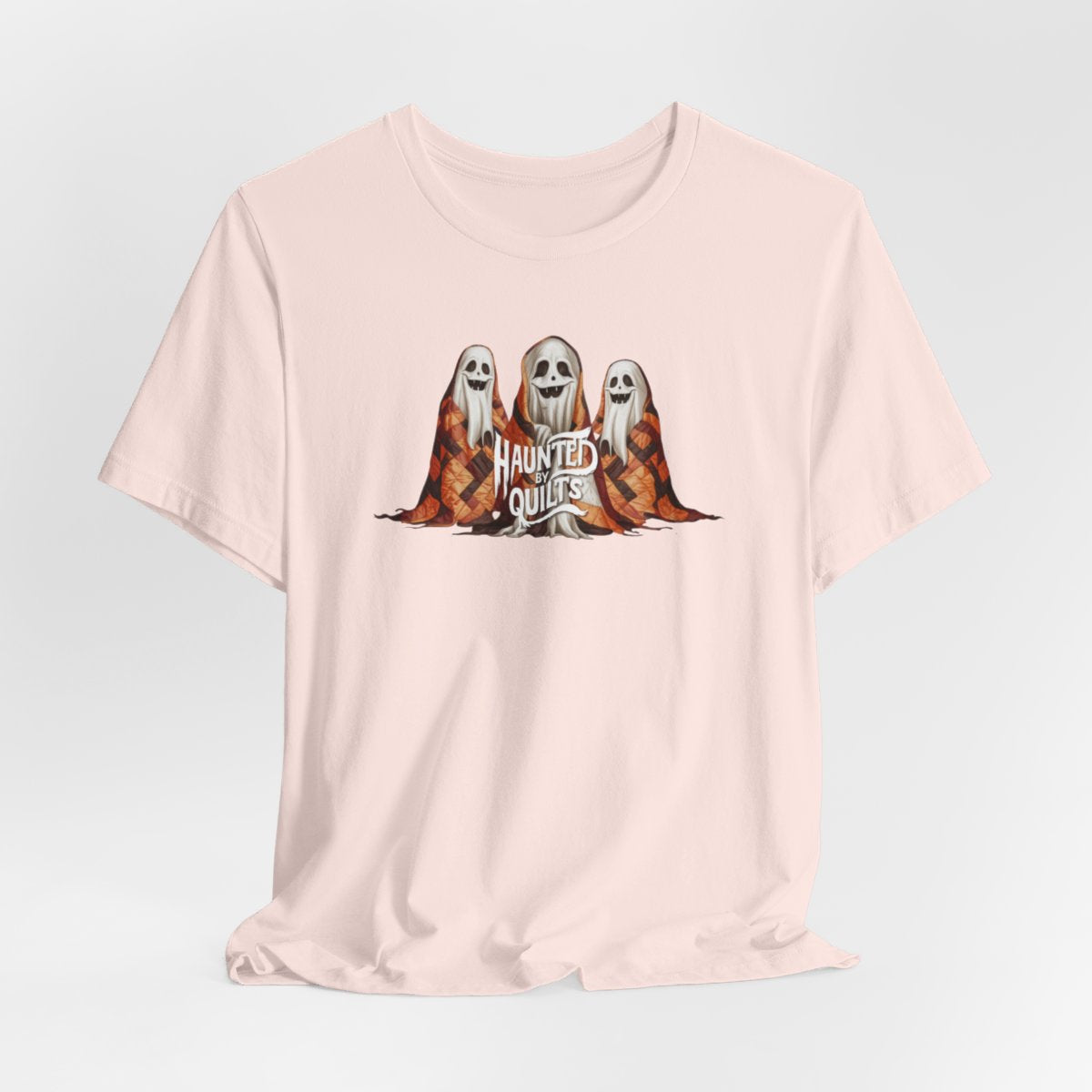 A Soft Pink T-Shirt of three smiling cartoon ghosts covered in colorful patchwork quilts. The quilts feature orange, brown, and black patterns. Text below reads "HAUNTED BY QUILTS" in a spooky white font.
