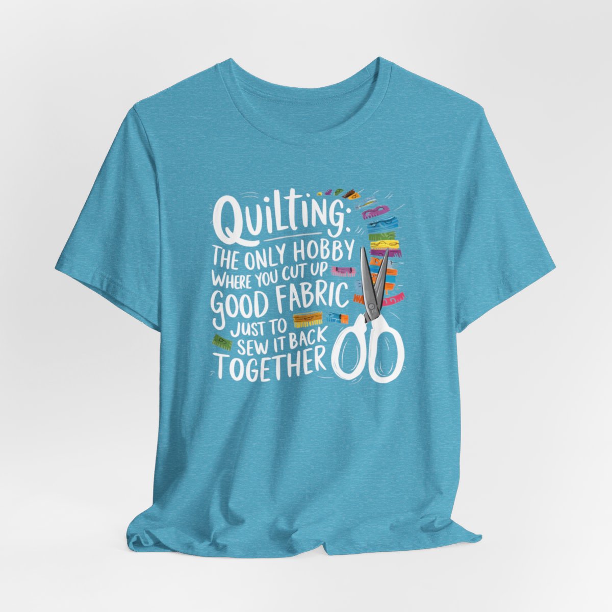 A Heather Aqua Quilting-themed T-Shirt with text "Quilting: The Only Hobby Where You Cut Up Good Fabric Just to Sew it Back Together" surrounded by colorful fabric scraps and scissors.