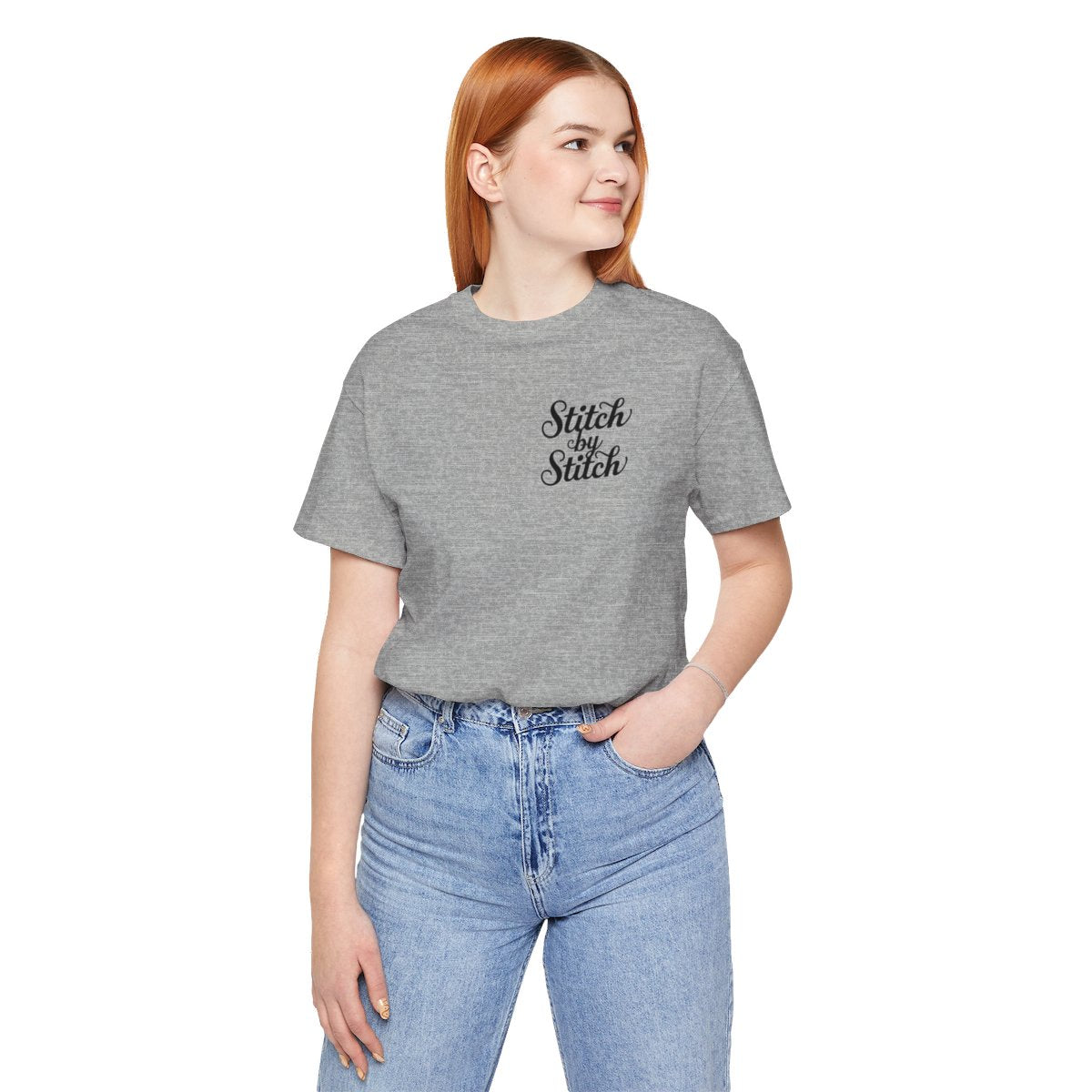 An Athletic Heather T-Shirt featuring the phrase "Stitch by Stitch". The word "Stitch" is written twice in a flowing script font, while "by" is centered between them in a smaller, sans-serif font. The design has a clean, minimalist aesthetic.