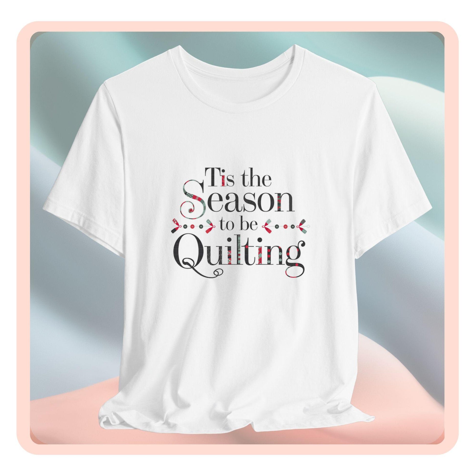 A festive t-shirt featuring the phrase "'Tis the Season to be Quilting," with playful sewing-inspired elements like needles, buttons, and fabric patterns integrated into the typography.