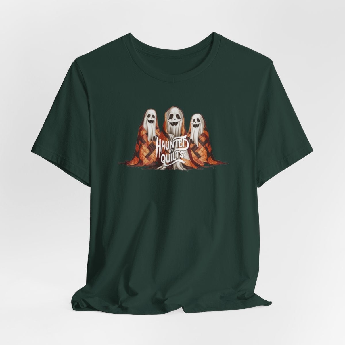 A Forest T-Shirt of three smiling cartoon ghosts covered in colorful patchwork quilts. The quilts feature orange, brown, and black patterns. Text below reads "HAUNTED BY QUILTS" in a spooky white font.