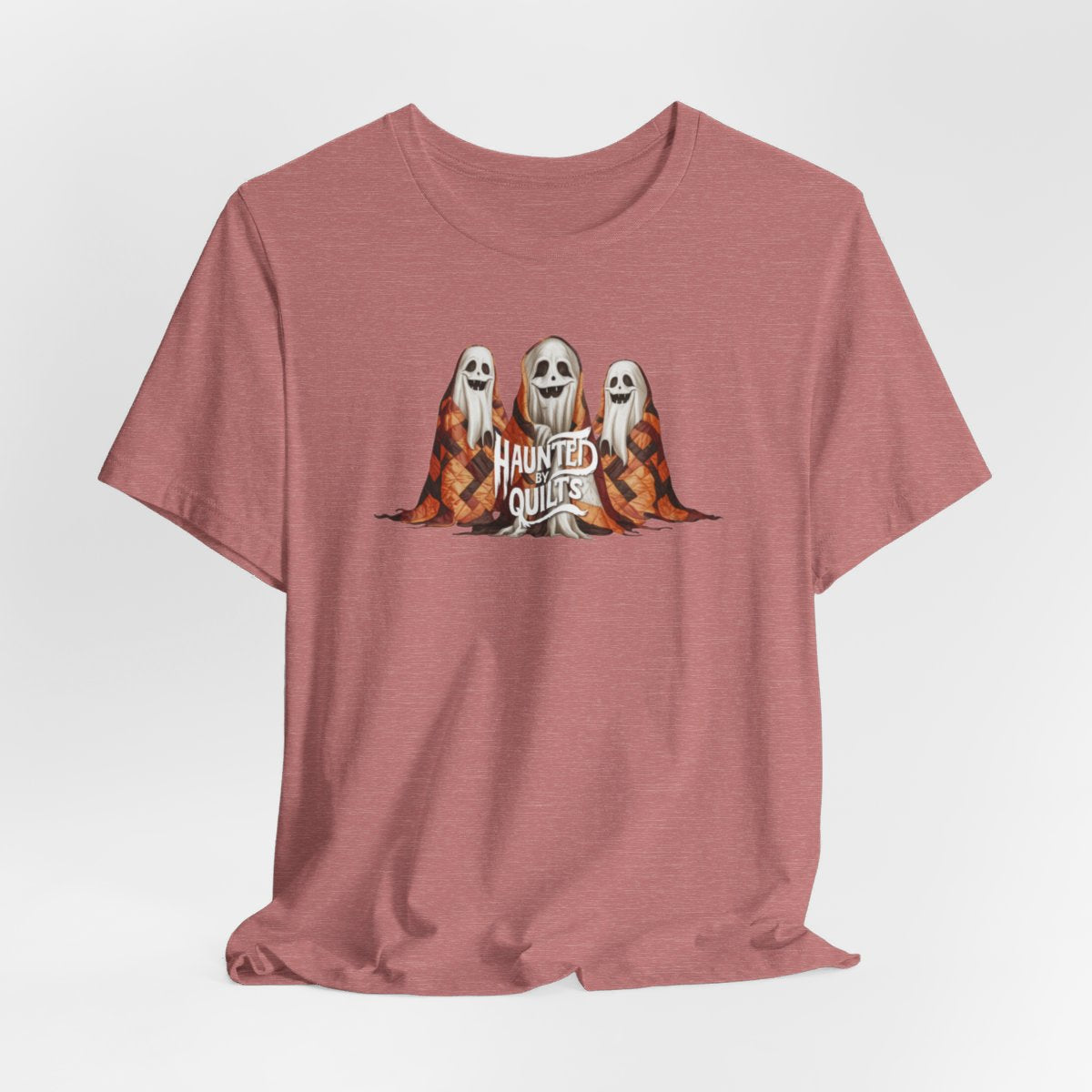 A Heather Mauve T-Shirt of three smiling cartoon ghosts covered in colorful patchwork quilts. The quilts feature orange, brown, and black patterns. Text below reads "HAUNTED BY QUILTS" in a spooky white font.