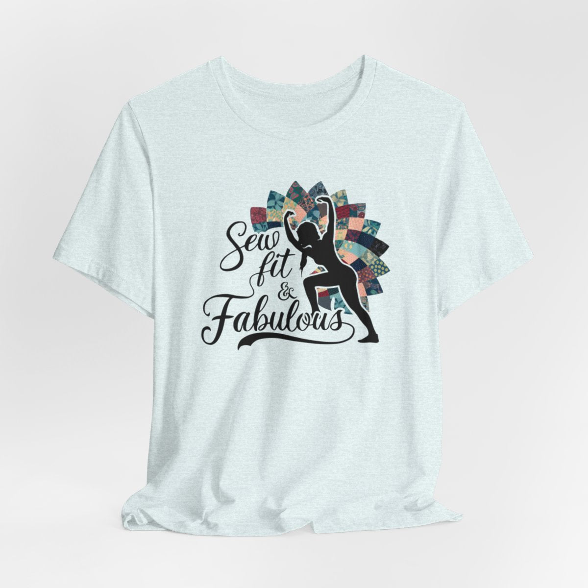 A Heather Ice Blue T-Shirt featuring a black silhouette of woman with raised arms against colorful patchwork background. Text reads "Sew fit & Fabulous" in stylized script.