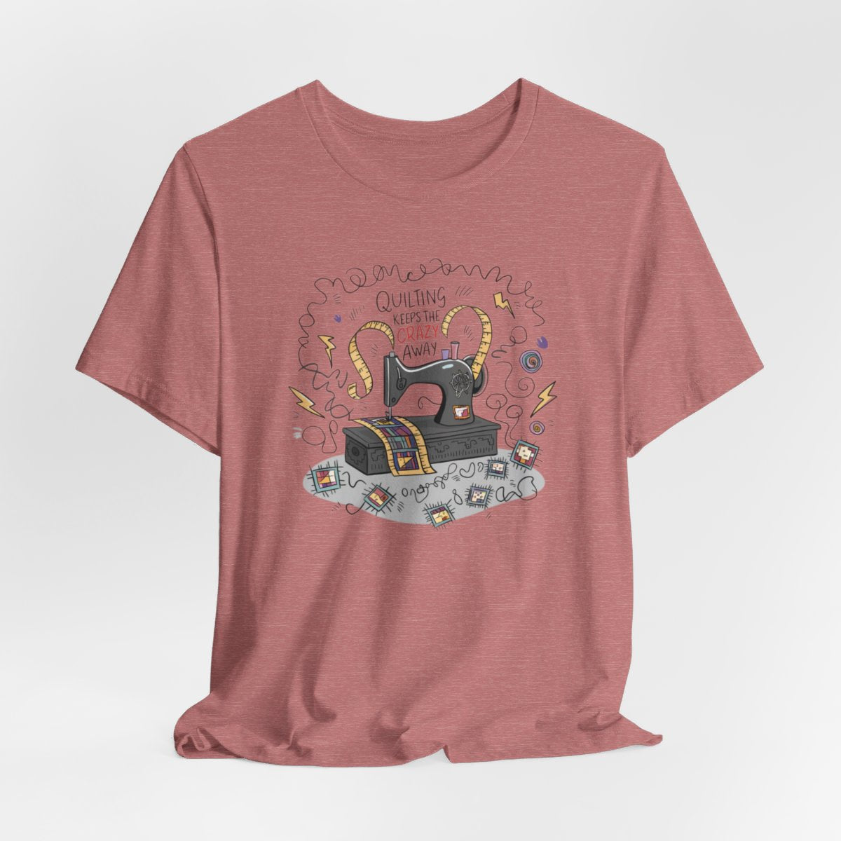 A Heather Mauve T-Shirt with a vintage sewing machine, measuring tape heart, and text "Quilting Keeps the Crazy Away" amidst swirls and quilting motifs.