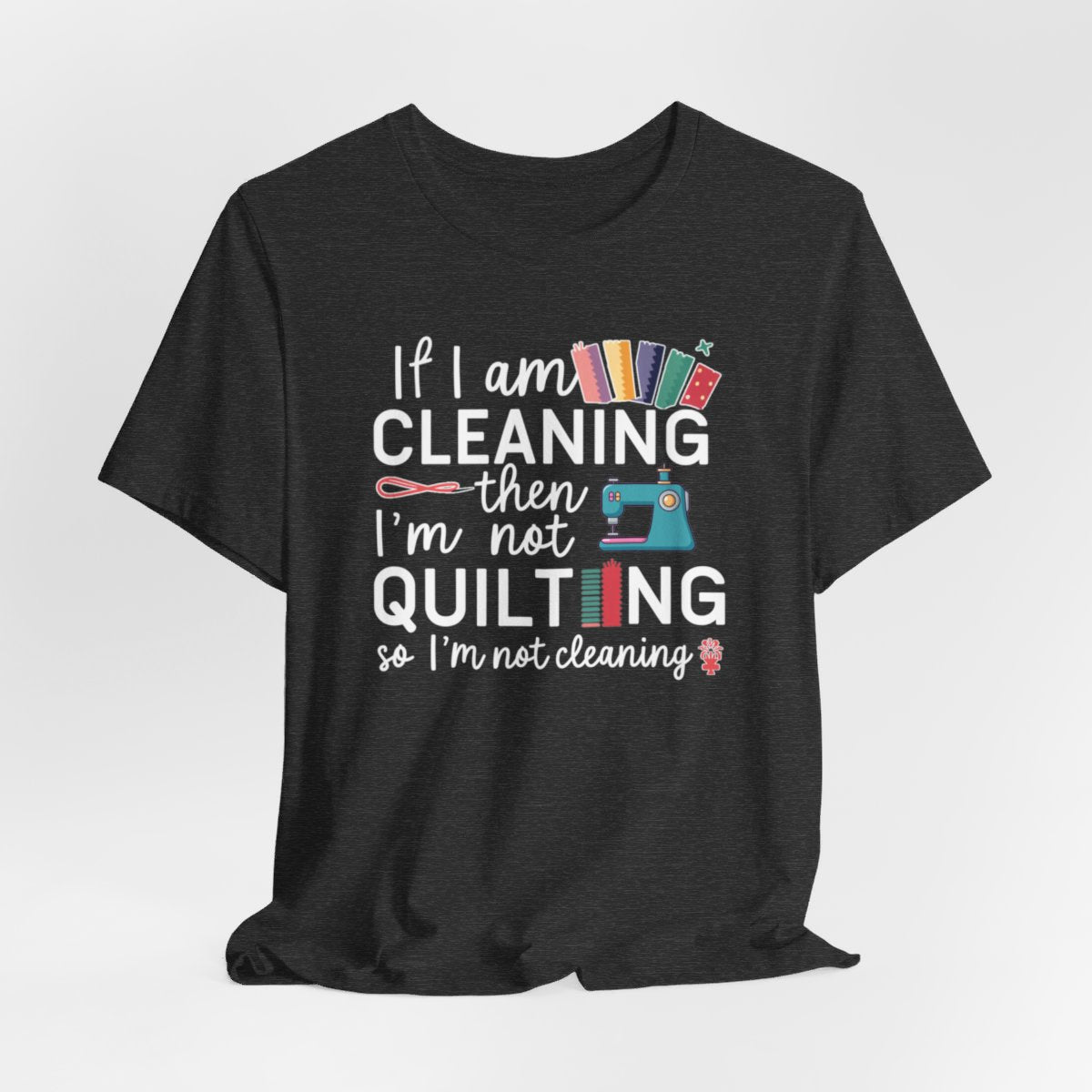 A Dark Grey Heather funny quilting T-Shirt with the phrase If I am Cleaning then I'm not Quilting in a bold and cheerful font with images of quilting icons and supplies