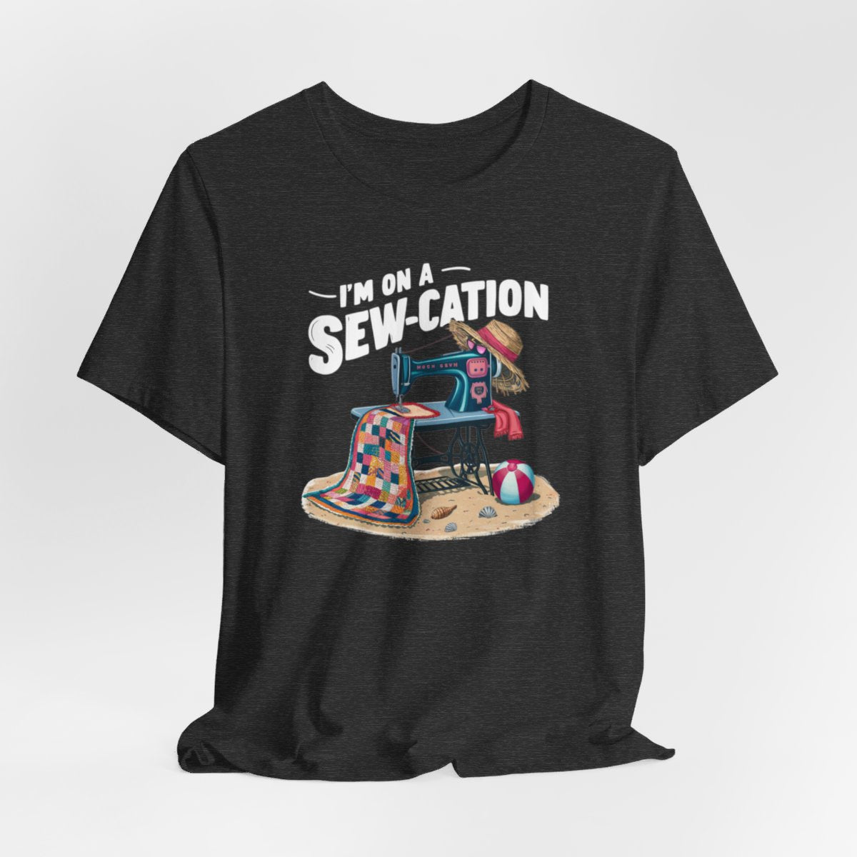 A Dark Grey Heather fun Quilting T-shirt of a vintage sewing machine on a beach with a colorful quilt, sun hat, and beach ball. Text above reads "I'm on a Sew-cation" in bold letters.