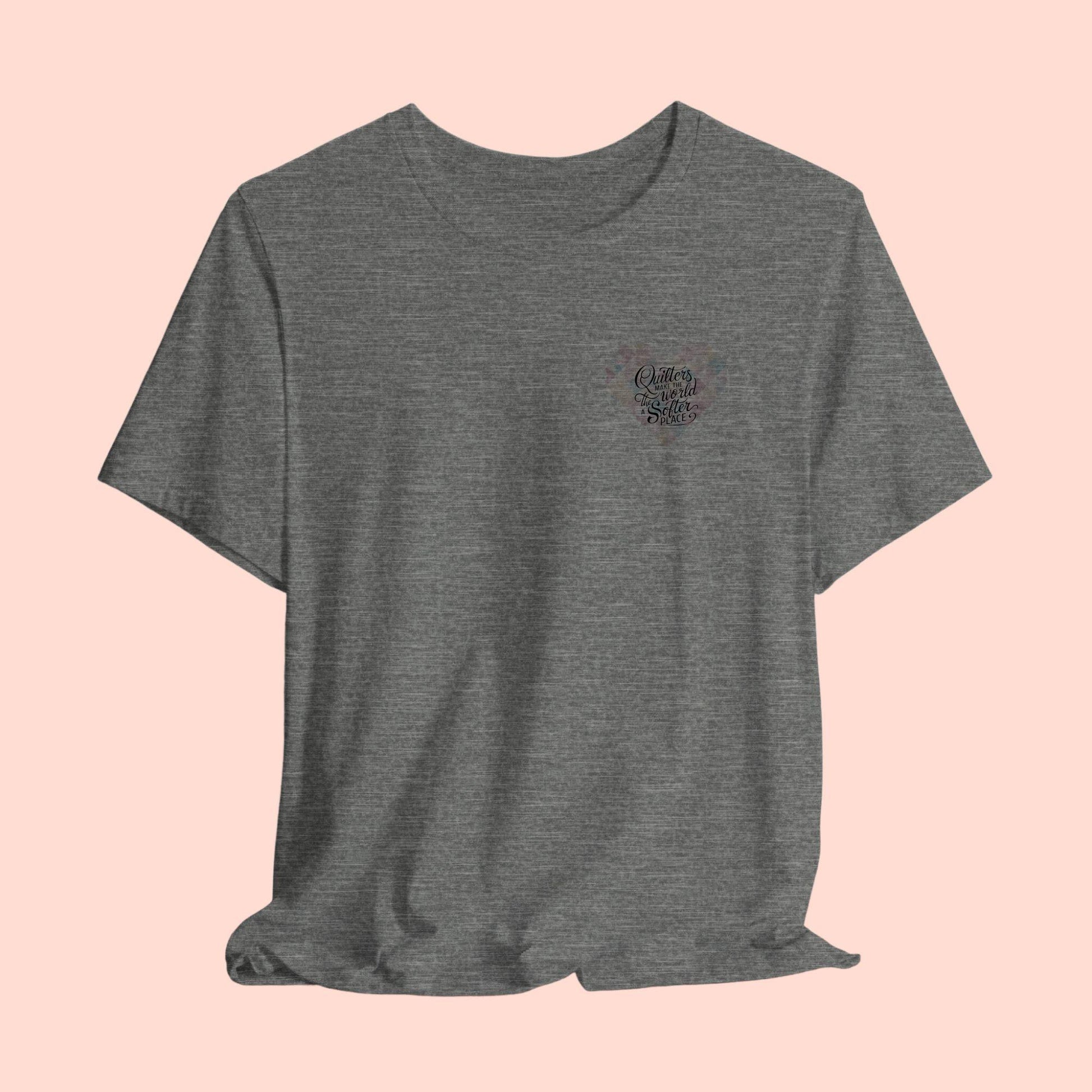 A T-shirt showing a large heart-shaped quilt pattern in pastel colors of pink, blue, and cream. Black text overlaid on the heart reads "Quilters make the world a softer place" in a mix of script and sans-serif fonts. The design combines geometric quilt patterns with an uplifting message about quilting.