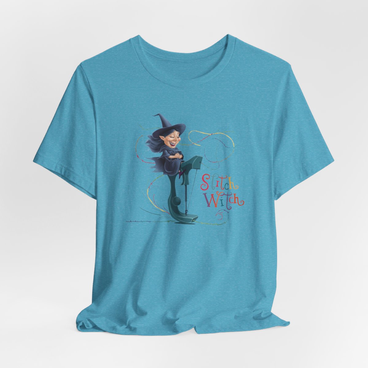 A Heather Aqua T-shirt with a cartoon illustration of a smiling witch with pointed hat riding a teal sewing machine. Colorful stitches swirl around her in the air. Text reads "Stitch Witch" in playful, multicolored letters.