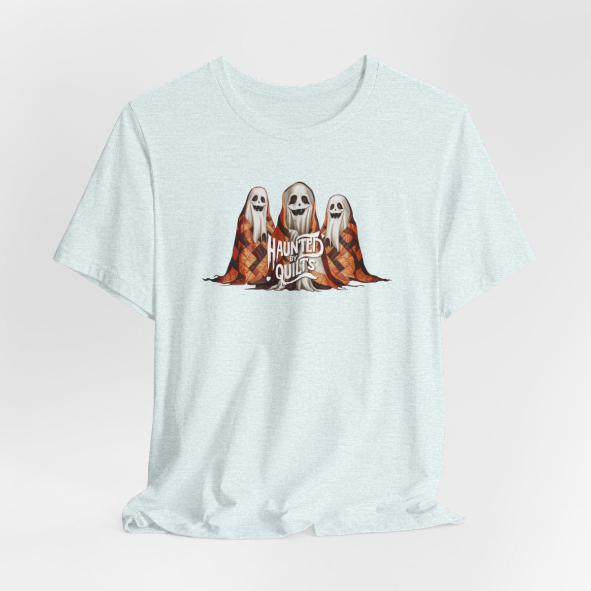 A Heather Ice Blue T-Shirt of three smiling cartoon ghosts covered in colorful patchwork quilts. The quilts feature orange, brown, and black patterns. Text below reads "HAUNTED BY QUILTS" in a spooky white font.