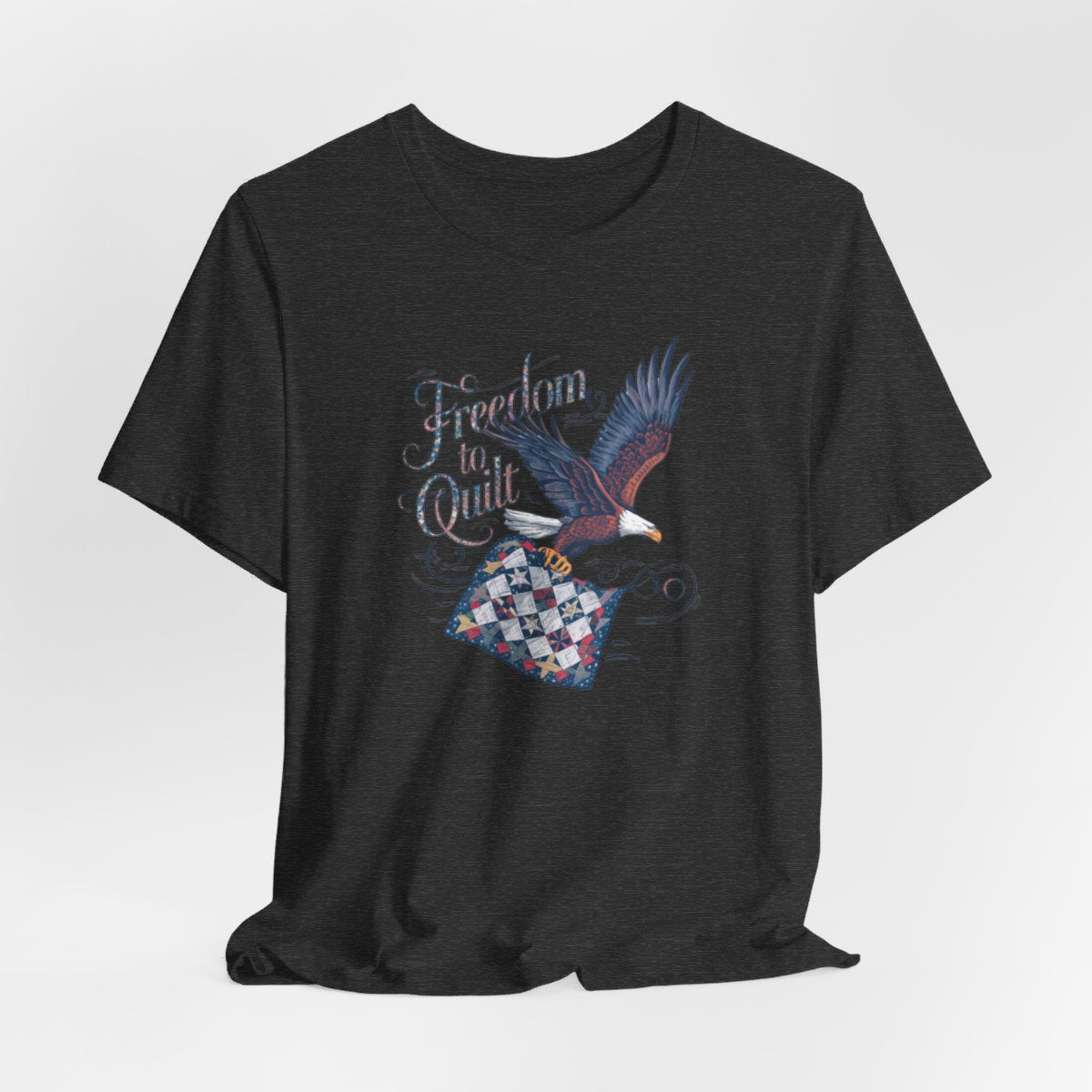 A Dark Grey Heather T-shirt with detailed illustration of a bald eagle in flight, carrying a patchwork quilt. Text reads "Freedom to Quilt" in an ornate, patriotic font.