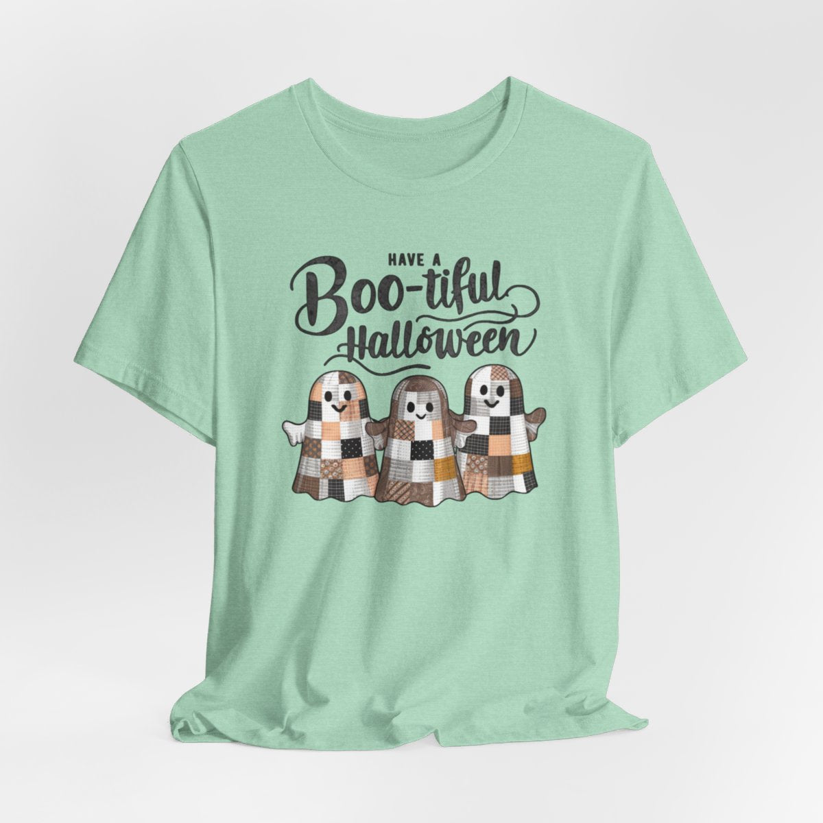 A Heather Mint Halloween Quilting T-Shirt featuring three smiling patchwork ghosts made of various quilt patterns in autumn colors. Text reads "Have a Boo-tiful Halloween" in stylized lettering. Perfect for quilters who love Halloween.