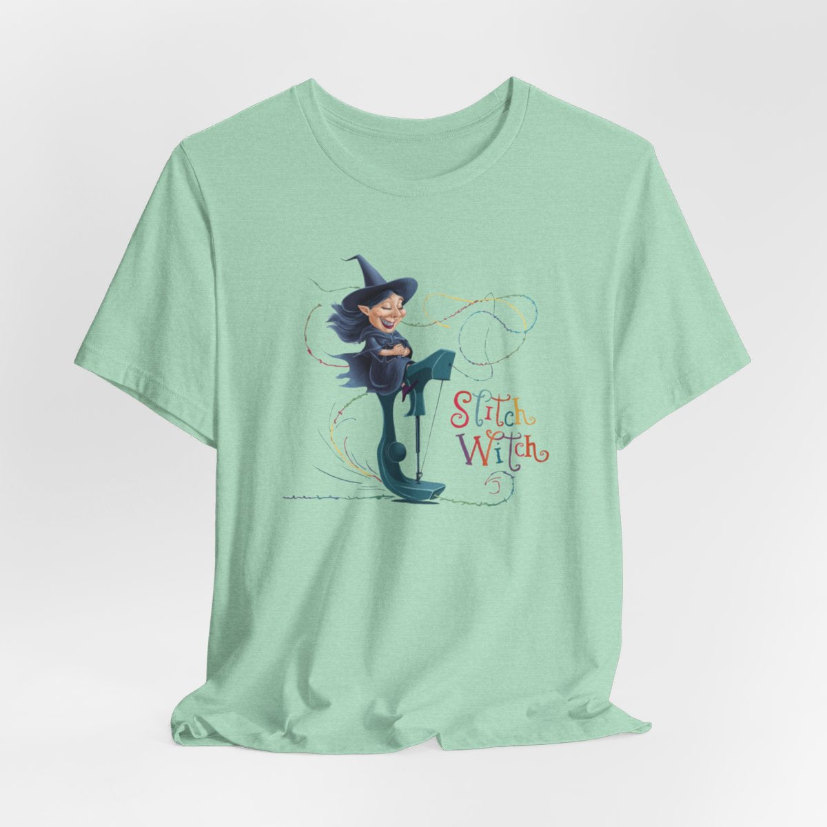 A Heather Mint T-shirt with a cartoon illustration of a smiling witch with pointed hat riding a teal sewing machine. Colorful stitches swirl around her in the air. Text reads "Stitch Witch" in playful, multicolored letters.