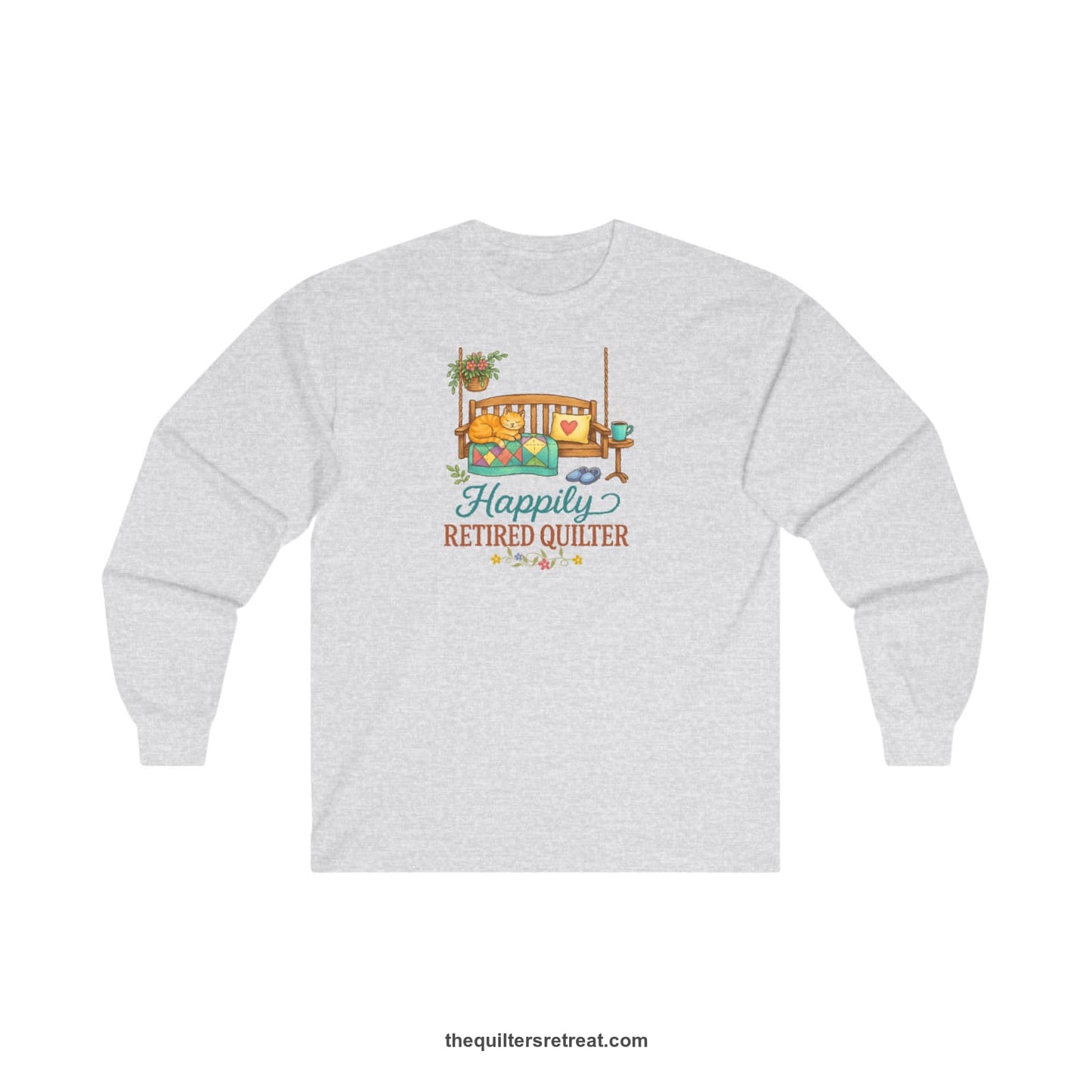 A Ash Long Sleeve T-Shirt with a hanging bench with cozy cat and quilt, surrounded by plants, "Happily Retired Quilter" text