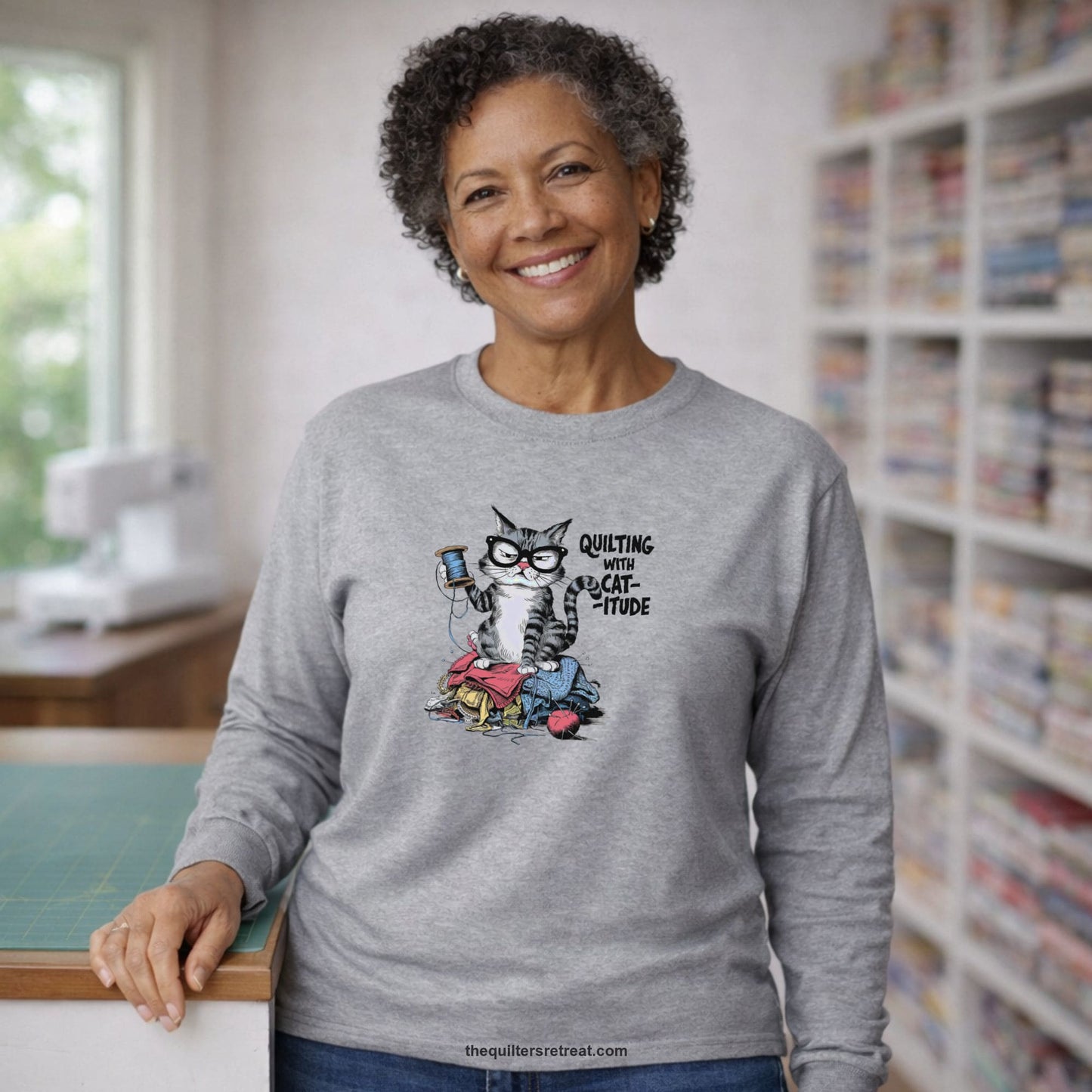 A Long Sleeve T-Shirt with a grumpy cat holding thread spool atop fabric pile with "Quilting with Cat-itude" text