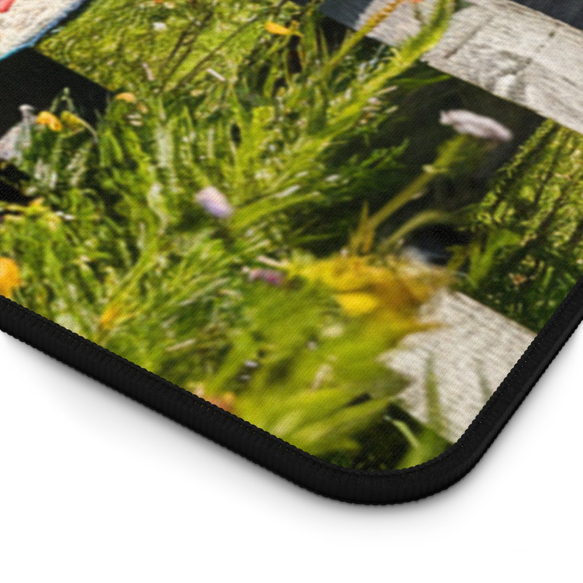 A quilting inspired Desk pad featuring a colorful patchwork quilt draped over a weathered wooden fence. Wildflowers in yellow, purple, and white surround the fence, with a blue sky and fluffy clouds above. Perfect gift for quilters.