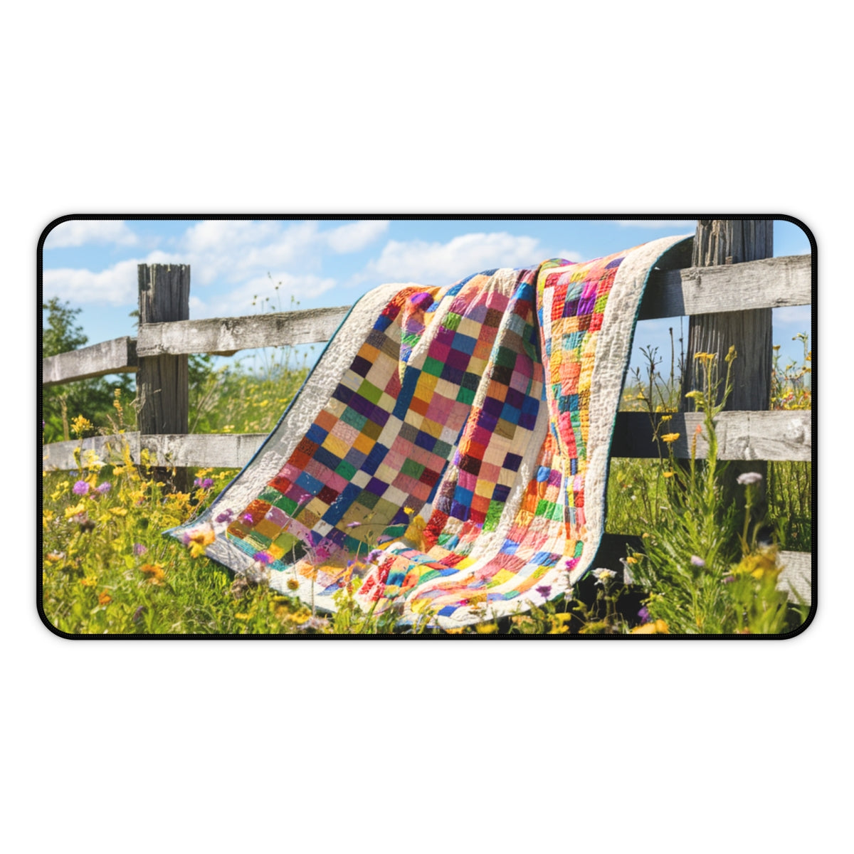 A quilting inspired Desk pad featuring a colorful patchwork quilt draped over a weathered wooden fence. Wildflowers in yellow, purple, and white surround the fence, with a blue sky and fluffy clouds above. Perfect gift for quilters.