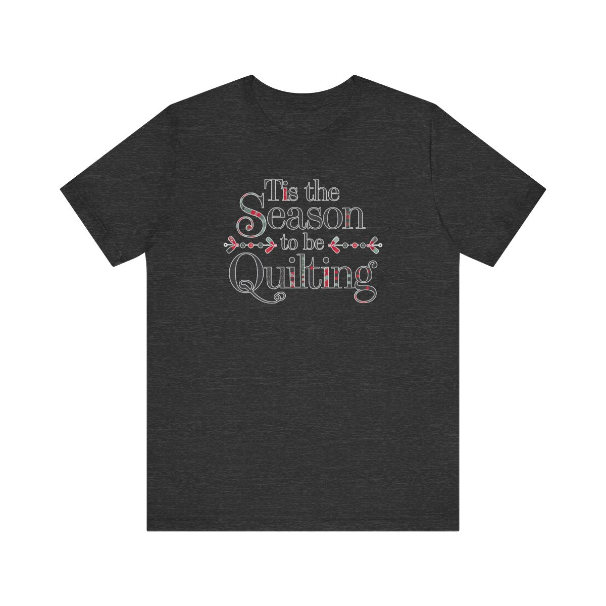 A Dark Grey Heather festive t-shirt featuring the phrase "'Tis the Season to be Quilting," with playful sewing-inspired elements like needles, buttons, and fabric patterns integrated into the typography.