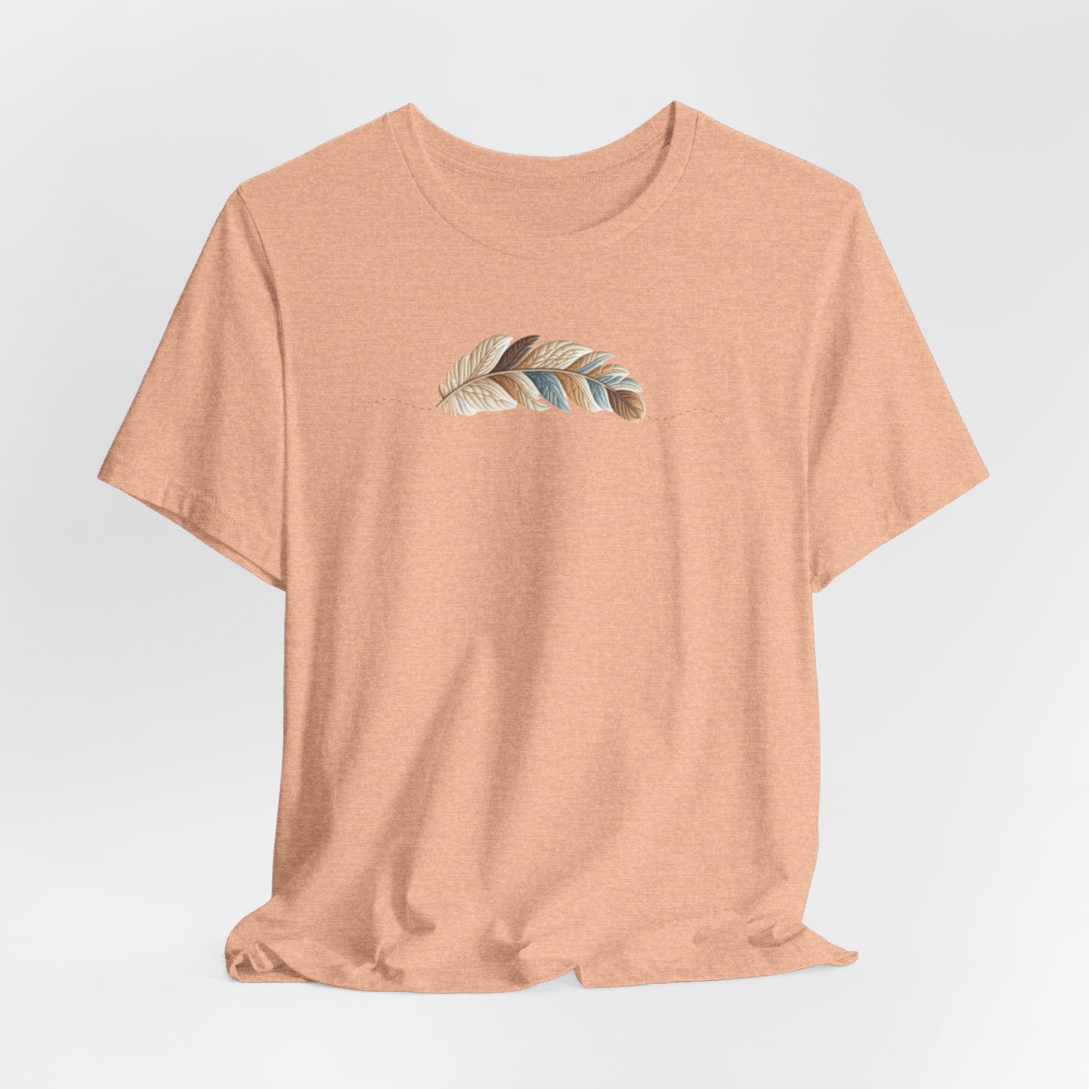 A Heather Peach T-Shirt with an illustration of a curving feather simulating patterned sections, resembling a patchwork quilt. The feather features a mix of floral, polka dot, and geometric designs in shades of blue, brown, and pink, with a dotted line tracing its path.