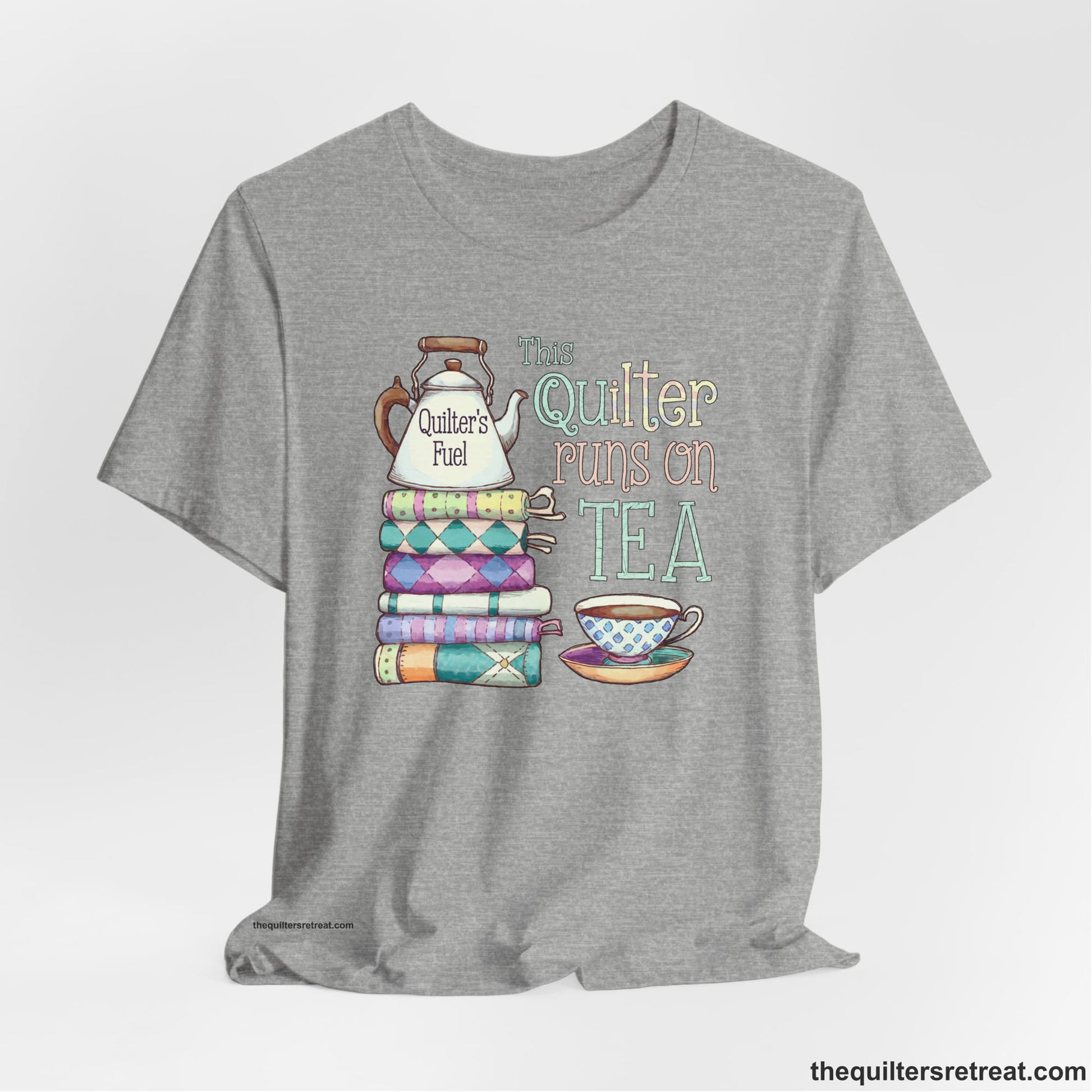 A Athletic Heather Short Sleeve T-Shirt with a stack of colorful quilts with teapot and cup, whimsical "This Quilter Runs on Tea" text
