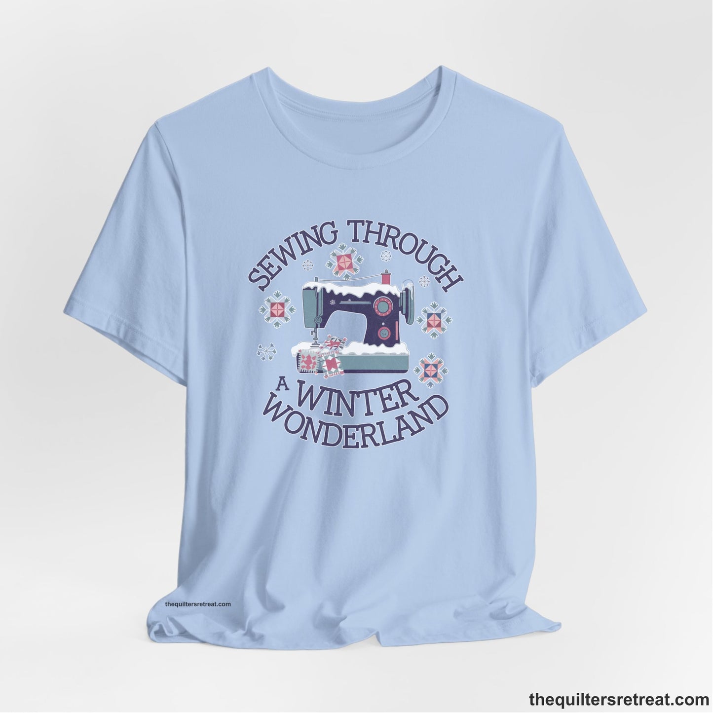 A Baby Blue Short Sleeve T-Shirt with a sewing machine covered in snow with festive quilting elements and "Sewing Through a Winter Wonderland" text