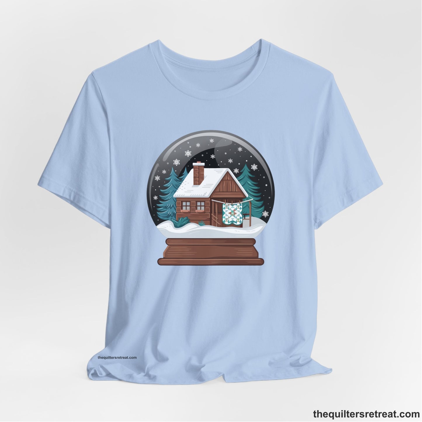 A Baby Blue Short Sleeve T-Shirt with a cabin in a snow globe with snowflakes and trees, in a cozy rustic style