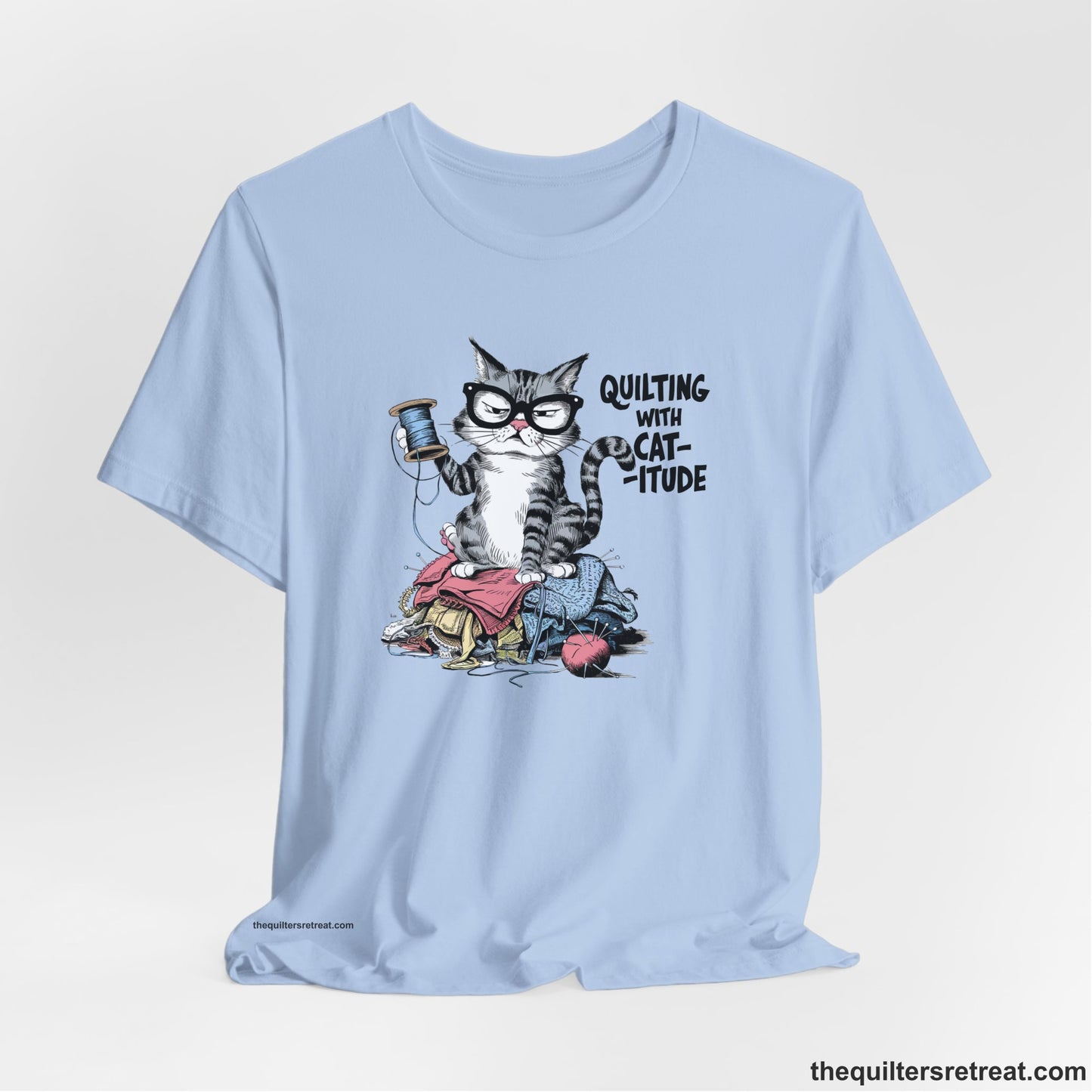 A Baby Blue Short Sleeve T-Shirt with a grumpy cat holding thread spool atop fabric pile with "Quilting with Cat-itude" text