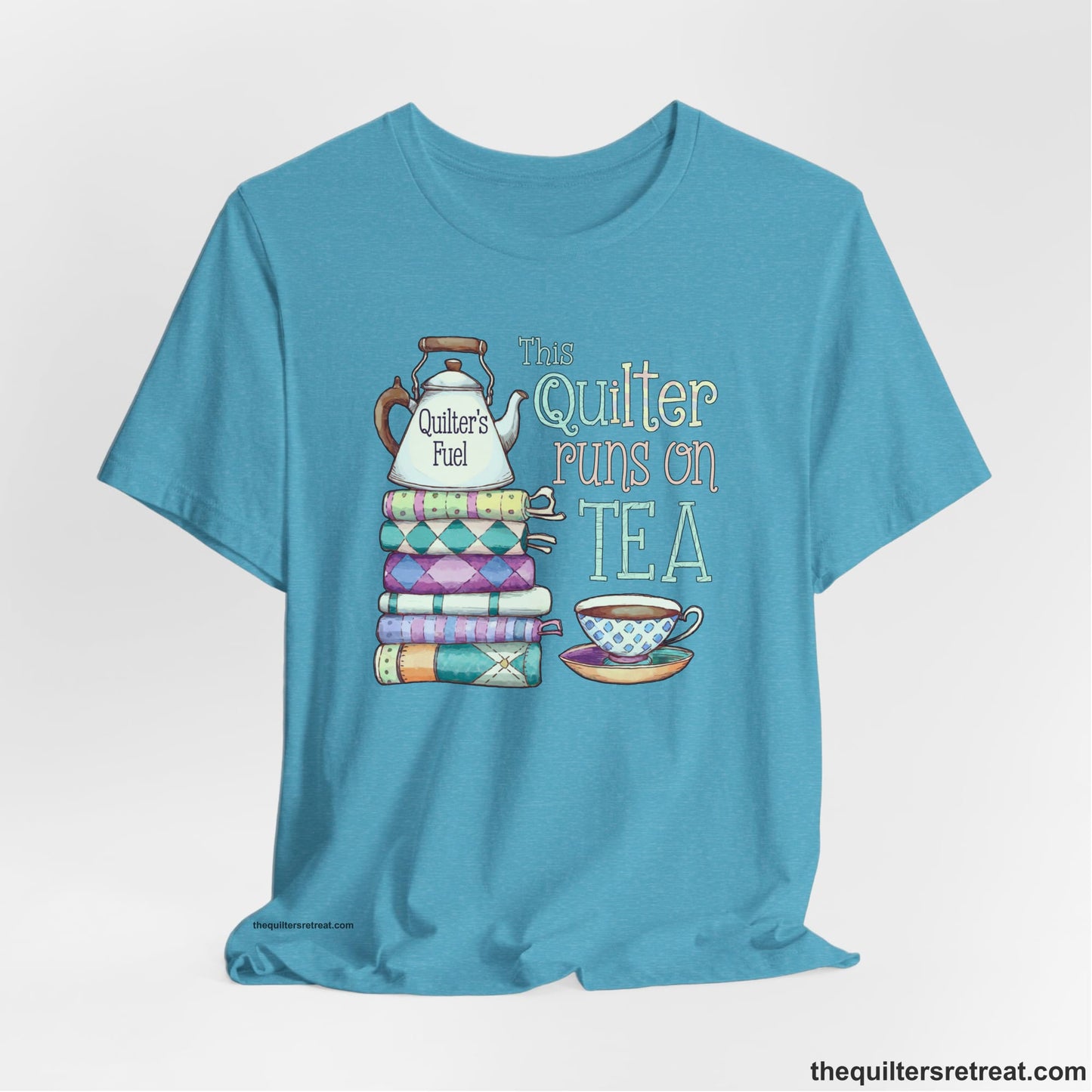 A Heather Aqua Short Sleeve T-Shirt with a stack of colorful quilts with teapot and cup, whimsical "This Quilter Runs on Tea" text