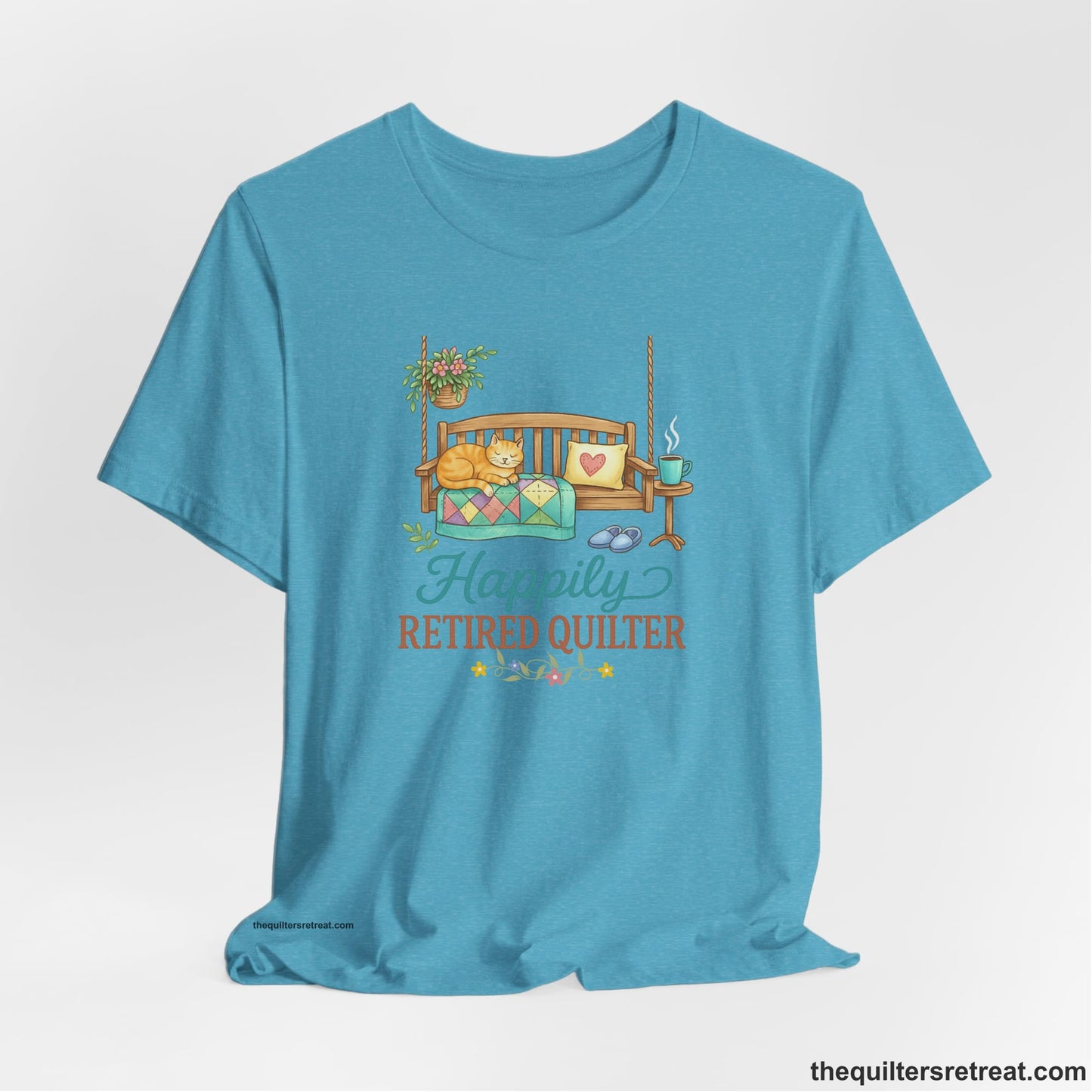 A Heather Aqua Short Sleeve T-Shirt with a hanging bench with cozy cat and quilt, surrounded by plants, "Happily Retired Quilter" text