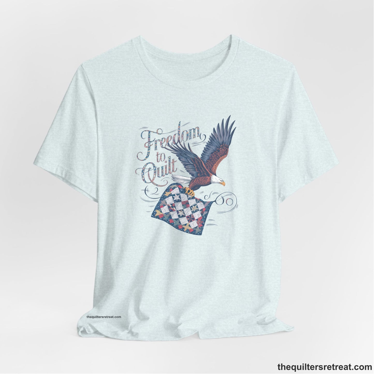 A Heather Ice Blue Short Sleeve T-Shirt with a freedom to quilt text with an eagle carrying a colorful patchwork quilt in vintage style