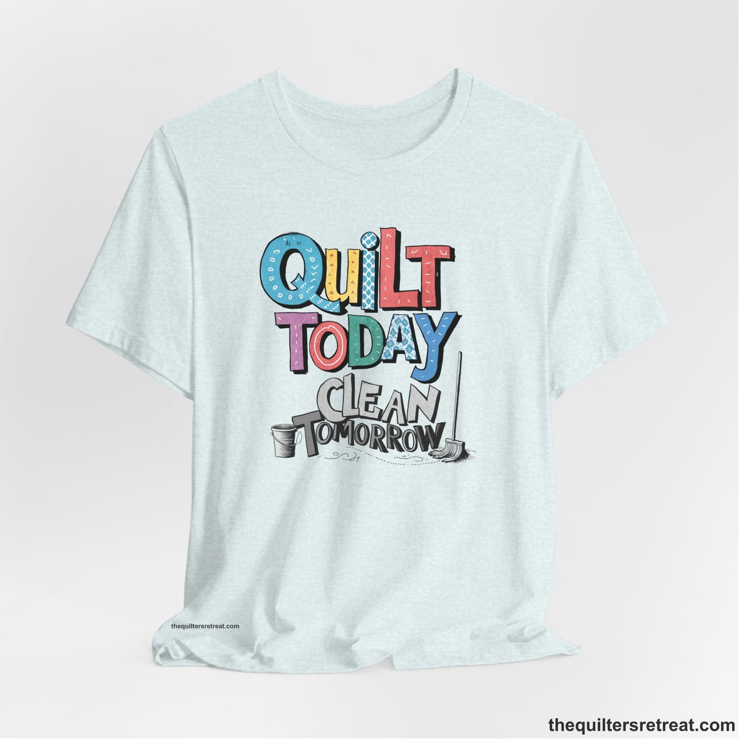 A Heather Ice Blue Short Sleeve T-Shirt with a playful "Quilt Today Clean Tomorrow" text with colorful patterns and cleaning tools illustration