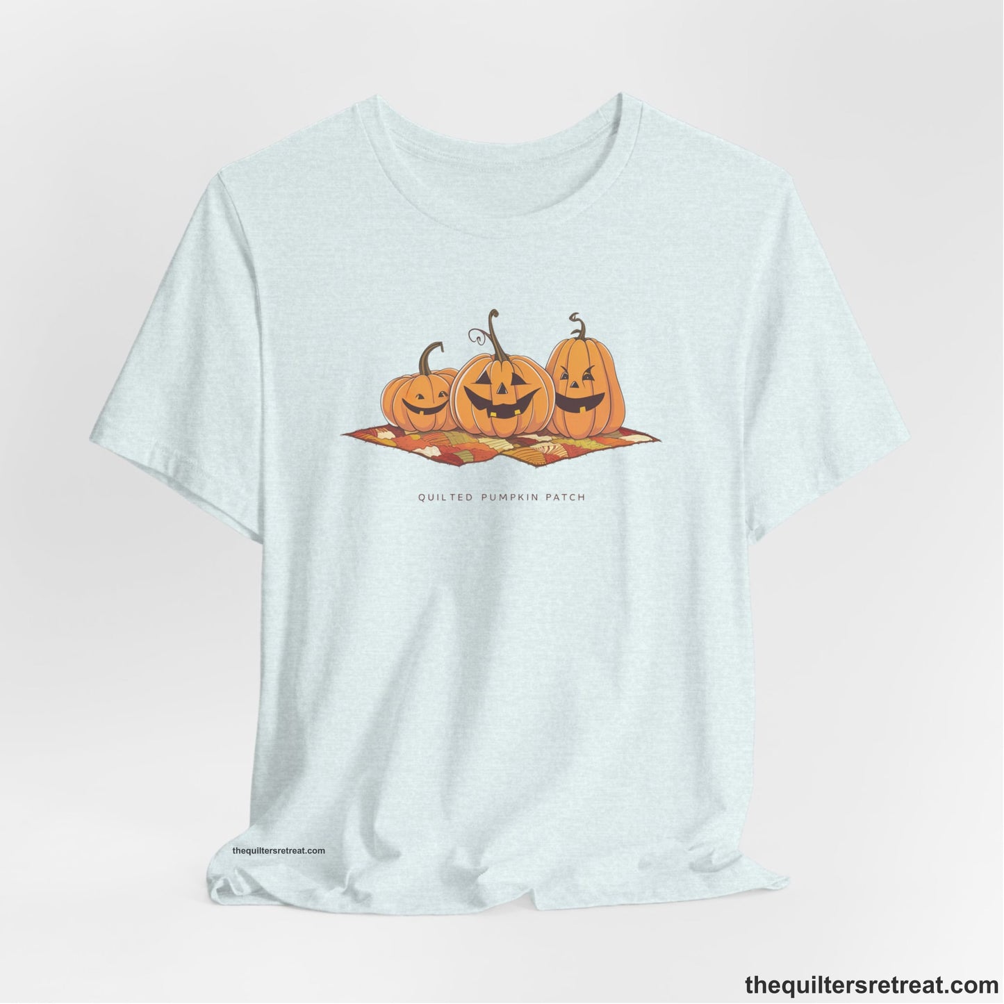 A Heather Ice Blue Short Sleeve T-Shirt with a playful jack-o'-lantern trio on a quilt with "Quilted Pumpkin Patch" text