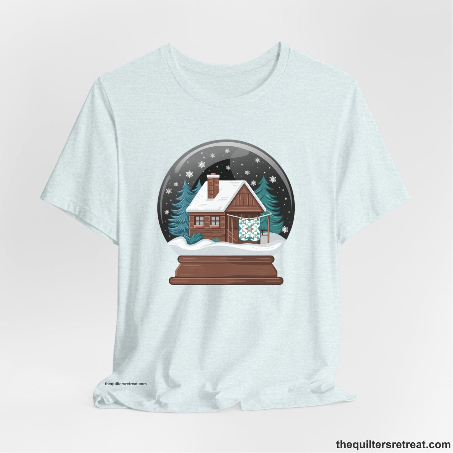 A Heather Ice Blue Short Sleeve T-Shirt with a cabin in a snow globe with snowflakes and trees, in a cozy rustic style