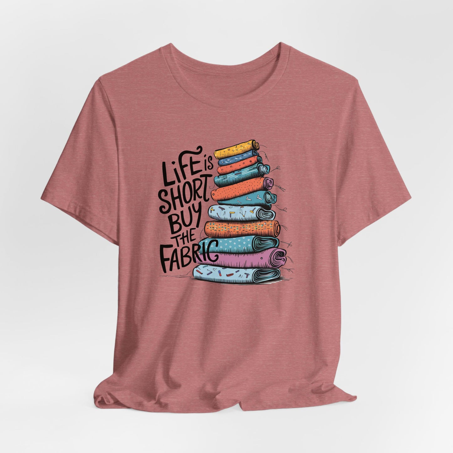 A Heather Mauve Short Sleeve T-Shirt with a illustration of colorful fabric rolls with bold "Life is Short Buy the Fabric" text
