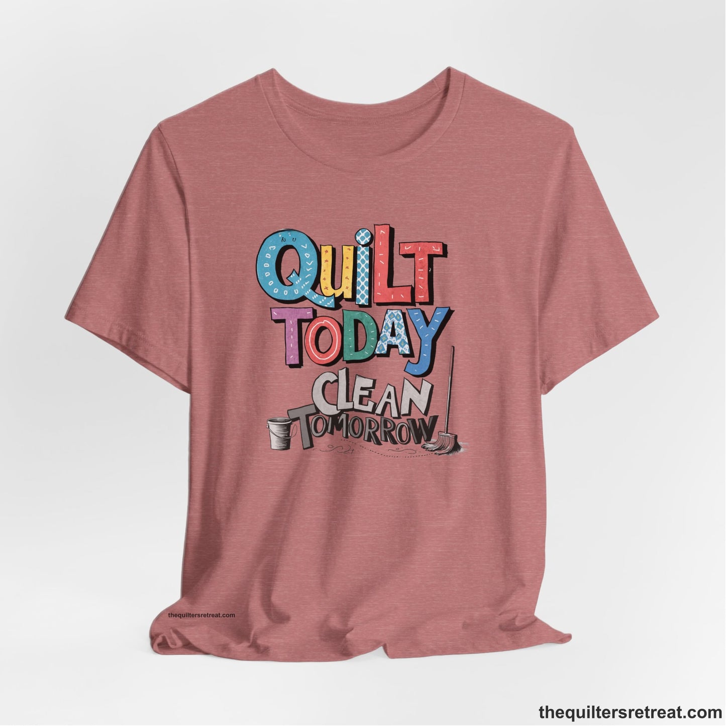 A Heather Mauve Short Sleeve T-Shirt with a playful "Quilt Today Clean Tomorrow" text with colorful patterns and cleaning tools illustration