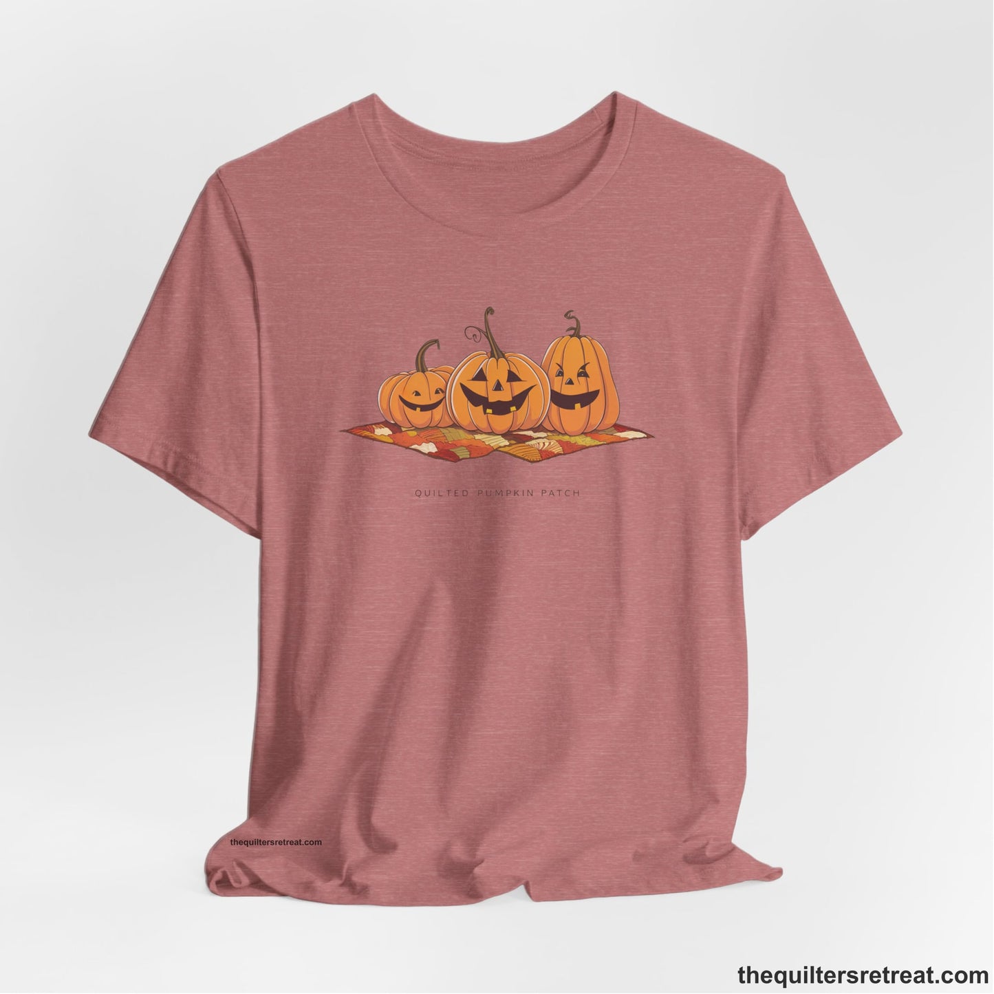 A Heather Mauve Short Sleeve T-Shirt with a playful jack-o'-lantern trio on a quilt with "Quilted Pumpkin Patch" text