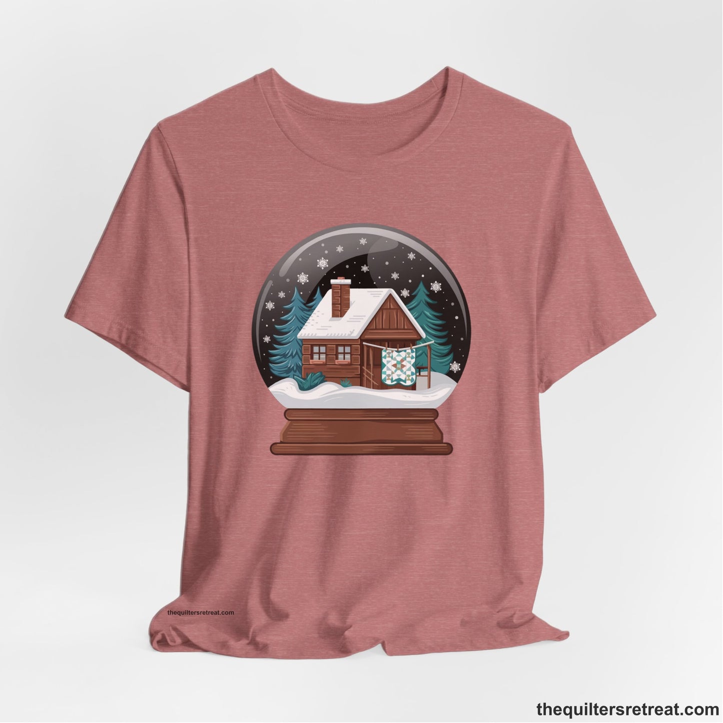 A Heather Mauve Short Sleeve T-Shirt with a cabin in a snow globe with snowflakes and trees, in a cozy rustic style