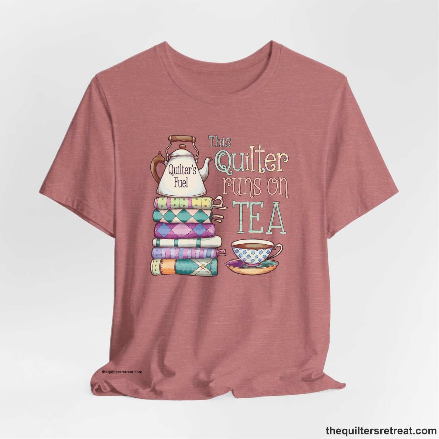 A Heather Mauve Short Sleeve T-Shirt with a stack of colorful quilts with teapot and cup, whimsical "This Quilter Runs on Tea" text