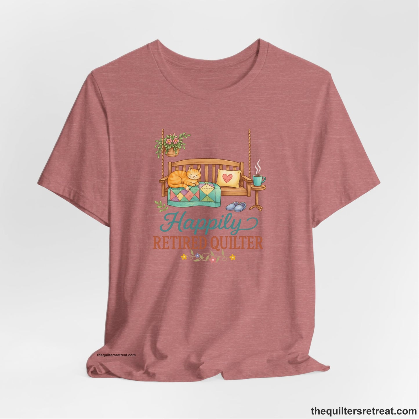 A Heather Mauve Short Sleeve T-Shirt with a hanging bench with cozy cat and quilt, surrounded by plants, "Happily Retired Quilter" text